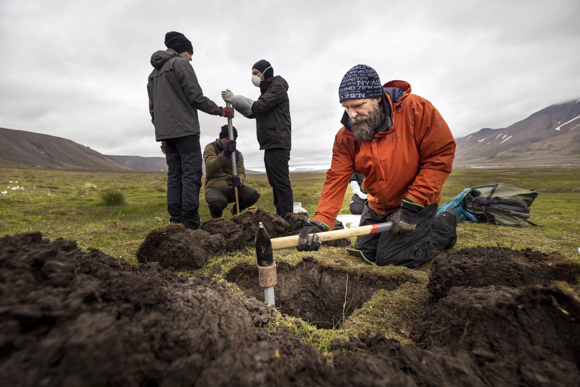 A researcher wearing a winter coat, hat, and gloves kneels next to a square hole, with the displaced soil piled around it. The researcher is using a long-handled hammer to hammer a metal cylinder into the hole. Behind him, three other researchers stand around another cylinder, which is about four feet long.